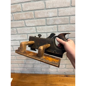 May include: A vintage woodworking plane featuring a dark metal body with a weathered finish, two light-colored wooden front handles, and a contoured wooden rear handle. The tool includes adjustable metal components and a flat base for smoothing wood.