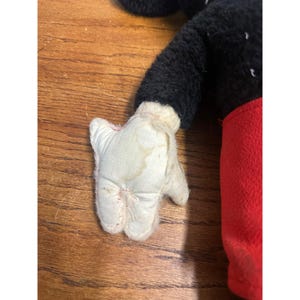 May include: Close-up of a vintage Mickey Mouse plush toy. The toy has a black body, red shorts, and white gloves. The plush shows signs of wear, with visible marks on the white glove.