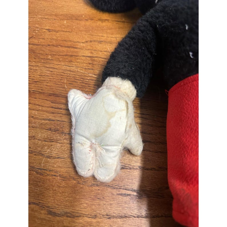 May include: Close-up of a vintage Mickey Mouse plush toy. The toy has a black body, red shorts, and white gloves. The plush shows signs of wear, with visible marks on the white glove.