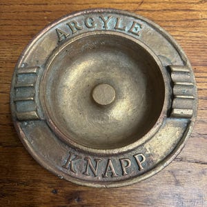 May include: A vintage brass ashtray with the words "ARGYLE" and "KNAPP" embossed on the rim. The ashtray has a circular design with a central depression and two built-in rests for cigarettes. The brass has a warm, aged patina.