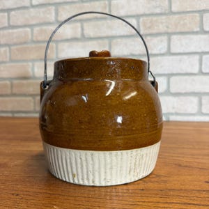 May include: A vintage brown ceramic jar with a lid and a metal handle. The jar has a white, ribbed base and a glossy finish. The handle arches over the top, connecting to the sides. The jar is likely used for storage.