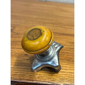 May include: A vintage-style door knob featuring a round, marbled amber-colored handle with a copper-toned coin embedded in the center. The base is constructed from a metallic, silver-colored material with a star-shaped mounting plate design.