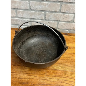 May include: A dark, cast iron cooking pot with a rounded shape and a metal handle. The pot has a textured surface and is sitting on a wooden surface. The background features a brick wall.