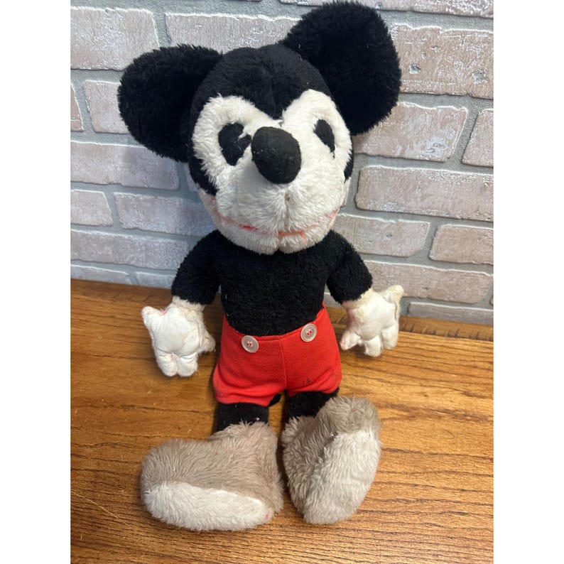 May include: Vintage Mickey Mouse soft toy. The toy has a black body, large black ears, white gloves, and red shorts. The face is white with a black nose and a red mouth. The plush is standing on a wooden surface.