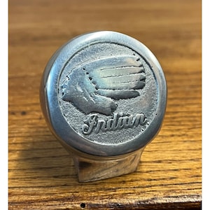 May include: A silver-colored, round knob with the word "Indian" in script. The knob features a detailed relief of a Native American profile with a feathered headdress. The knob has a curved base and is likely a decorative or functional item.
