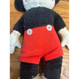 May include: A vintage Mickey Mouse soft toy, seen from the rear. The toy has a black body, red shorts with white buttons, white gloves, and a white and black head. It is placed on a wooden surface.