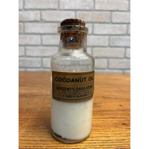 May include: A clear glass bottle with a cork stopper containing a white substance. The label reads COCOANUT OIL, SARGENT'S DRUG STORE, HERZ BROS. PROPRIETORS, 23 N. WABASH AVE. CHICAGO. The bottle has a cylindrical body and a narrow neck with a cork.