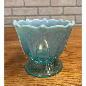 May include: A light blue, antique glass bowl with a pedestal base. The bowl has a scalloped rim and is decorated with a pattern of teardrop shapes and small dots. The glass has a slight iridescence.