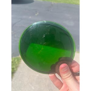 May include: A round, transparent, green glass object with a slightly raised edge. The object has some scratches and imperfections on its surface. The item is being held up in front of a bright, outdoor background.