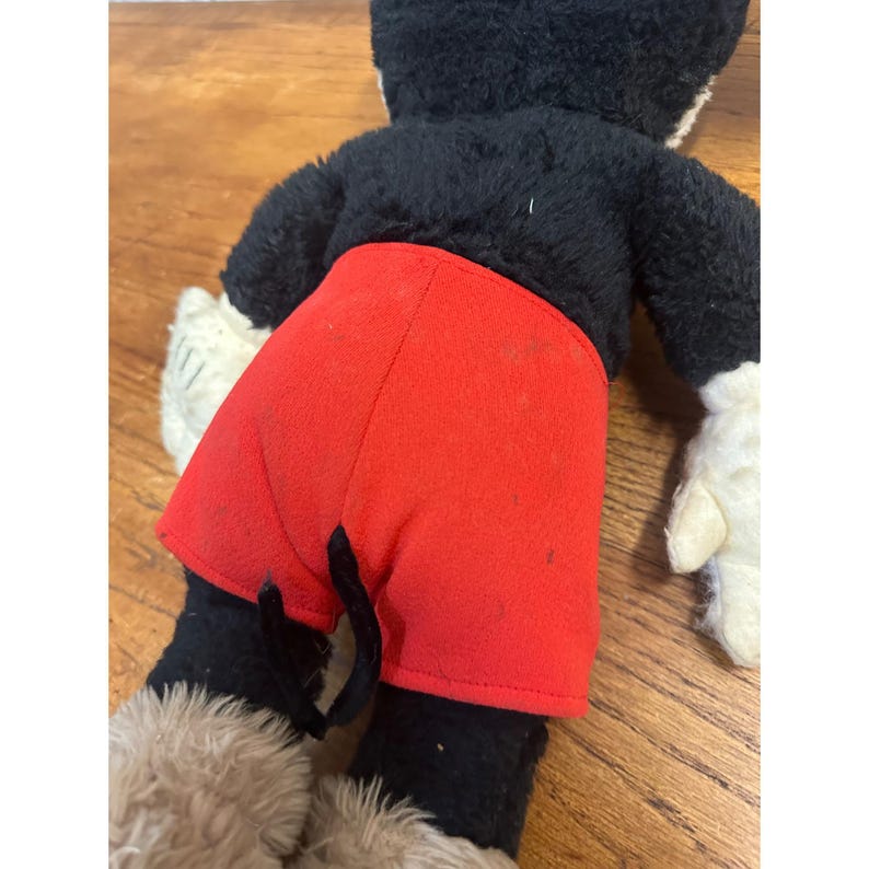 May include: A vintage Mickey Mouse soft toy, seen from the rear. The toy has a black body, red shorts, white gloves, and tan fur on the feet. The toy is placed on a wooden surface.