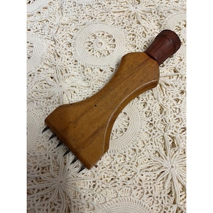 May include: A wooden meat tenderizer featuring a contoured handle and a dark brown rubberized cap at the top. The base of the tool is wide and flat, equipped with five sharp, pointed metal prongs extending from the bottom edge for piercing food.