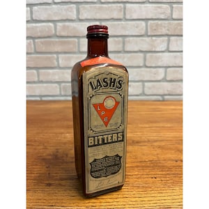 May include: A vintage brown glass bottle of Lash's Bitters with a red cap. The label features the brand name and the words "Natural Laxative." The bottle is rectangular with a detailed label and is set against a brick wall background.