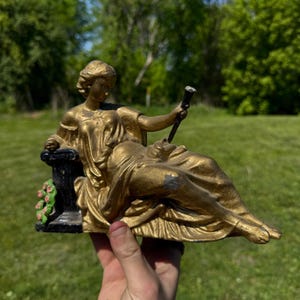 May include: A gold-colored statue of a reclining woman holding a staff. The statue is seated on a black pedestal with pink and green floral accents. The statue is held in a person's hand against a backdrop of green grass and trees.