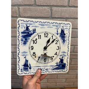 May include: Square, white wall clock with a blue and white windmill and sailboat design. The clock face has black numbers and hands, and a key is inserted. The clock is being held.