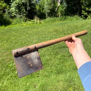 May include: A vintage hand tool featuring a long, cylindrical wooden handle attached to a wide, flat, rectangular metal blade. The metal blade shows signs of oxidation and surface rust, while the wooden handle is secured with two metal bolts.