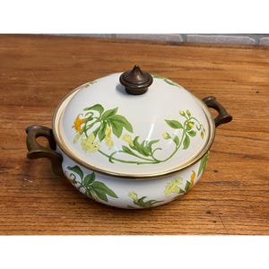 May include: White enamelware casserole dish with lid. The dish features a floral design with yellow flowers and green leaves. It has bronze-colored handles and a bronze knob on the lid. The dish is sitting on a wooden surface.