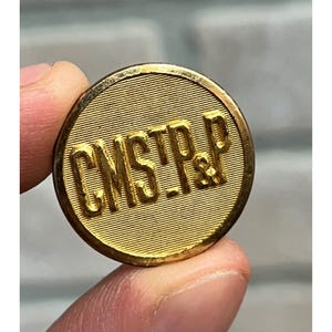 May include: A circular gold-colored metal button featuring a fine horizontal line texture across the surface. The letters CMST.P&P are embossed in a bold, raised font across the center of the button, creating a distinct contrast against the background.
