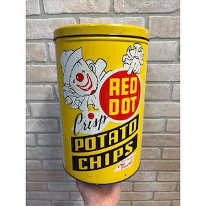 May include: A vintage yellow Red Dot Crisp Potato Chips tin. The tin features a clown graphic and the words "Red Dot Crisp Potato Chips" in bold lettering. The tin is cylindrical and appears to be made of metal.