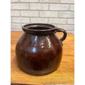 May include: A dark brown glazed ceramic pot with a wide, rounded body and a single side handle. The vessel features a smooth, reflective surface and a thick, circular rim opening at the top, showcasing a classic, utilitarian design with a deep, rich color.