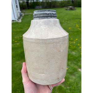 May include: A vintage stoneware jug with a light beige body and a dark gray, ribbed top. The jug has a cylindrical shape with a slightly flared neck. The overall aesthetic suggests a rustic, antique style, suitable for farmhouse decor.
