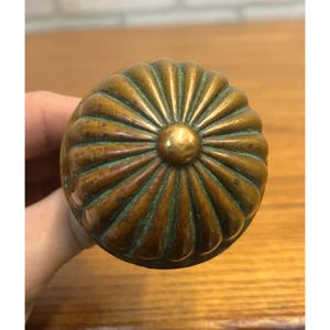 May include: A round, fluted brass door knob with a weathered, aged patina. The surface features a series of radiating ridges that extend from a central circular point to the outer edge, showing dark oxidation in the crevices of the metal.