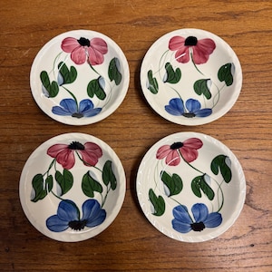 May include: Set of four small, round, white ceramic dishes. Each dish features a hand-painted floral design with pink and blue flowers, green leaves, and black centers. The dishes are arranged on a wooden surface.