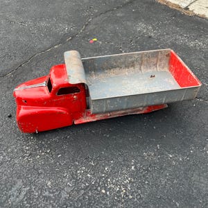 May include: A vintage red toy dump truck with a gray bed. The truck has a classic design with a curved hood and a roll-down bed. The truck is made of metal and appears to be well-used.