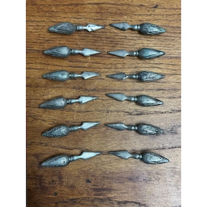 May include: A collection of twelve silver-toned metal decorative arrowheads, each with a textured, cone-shaped head and a pointed, symmetrical blade. The arrowheads are arranged on a wooden surface.