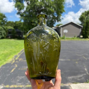 May include: An olive green glass flask featuring an embossed figure of a traveler carrying a bindle on a stick. The text FOR PIKES PEAK is molded into the glass above the figure. The bottle has a narrow neck and a rounded, flattened body shape.
