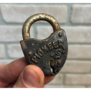 May include: An antique, weathered padlock with the word "PIONEER" embossed on the front. The lock is dark gray with a rusty, arched handle. The keyhole is visible at the bottom. The lock is likely made of metal and shows signs of age.