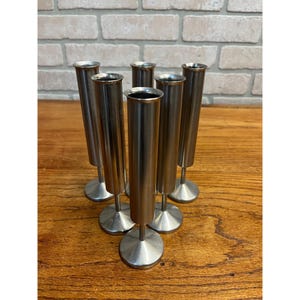 May include: A set of seven silver-colored metal bud vases. Each vase has a slender, cylindrical body and a circular base. The vases are arranged on a wooden surface, with a brick wall in the background.