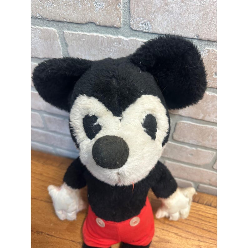May include: A vintage Mickey Mouse soft toy. The plush has a black body, large black ears, a white face and hands, and red shorts with white buttons. The toy is standing on a wooden surface.