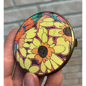 May include: A round, vintage compact mirror with a floral design. The compact has a brass-colored metal frame and a fabric top with a pattern of yellow sunflowers, orange and pink accents. The compact is held in a hand.