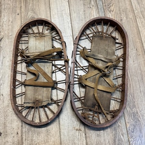 May include: Pair of vintage snowshoes. Each snowshoe has an oval wooden frame with a woven rope webbing. The footbeds are made of canvas and secured with brown straps. The snowshoes are a rustic brown color.