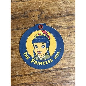 May include: A circular blue tag with a red metal eyelet at the top. The center features a yellow circle with an illustration of a cartoon character with dark hair, a headband with a red feather, and a white beaded necklace. The text reads THE PRINCESS says...