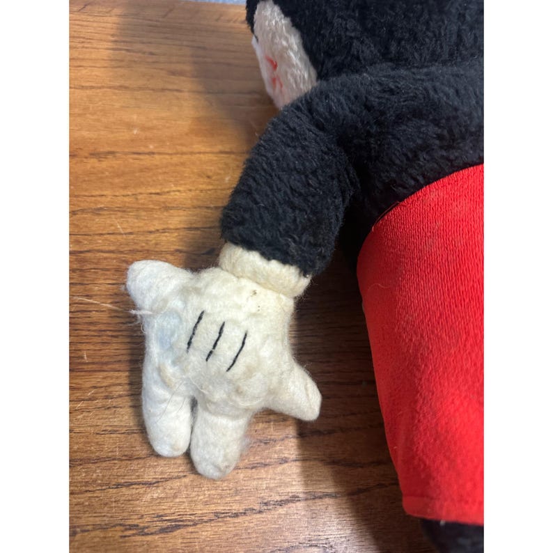 May include: A vintage Mickey Mouse plush toy. The plush has a black body, red shorts, and white gloves with three black lines on each hand. The toy's face is visible, with a white face and black ears.