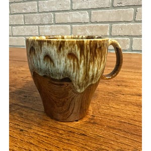 May include: A ceramic coffee mug with a brown and cream glaze. The mug has a rounded body and a handle. The glaze has a dripped effect, with the cream color at the top and the brown color at the bottom. The mug is sitting on a wooden surface.