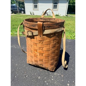 May include: A tall, cylindrical woven wood basket featuring a dark brown rim secured with metal rivets. The basket includes a tan canvas strap system with a metal buckle for carrying, designed with a flat base and a sturdy, open-top construction.