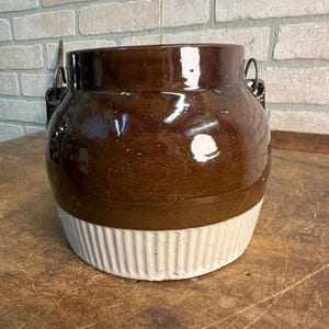 May include: A brown and white ceramic crock with a glossy finish. The upper portion is brown, while the lower section features a ribbed, off-white design. It has two metal handles and is likely used for storage or decoration.