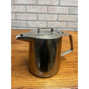 May include: A stainless steel pitcher with a lid and handle. The pitcher has a spout for pouring and is sitting on a wooden surface. The background is a brick wall.