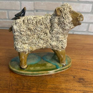 May include: Ceramic sheep figurine with a textured, off-white woolly coat and brown legs. A small black bird perches on its back. The sheep stands on a green and blue oval base, a unique decorative item.