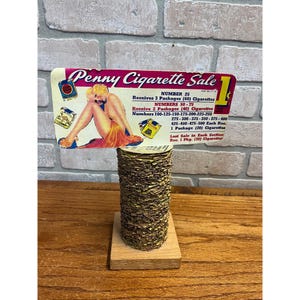 May include: A vintage Penny Cigarette Sale sign featuring a blonde woman in a yellow dress, mounted on a tall, cylindrical stack of small, thin, gold-colored paper tickets resting on a square wooden base. The sign lists various cigarette package rewards.