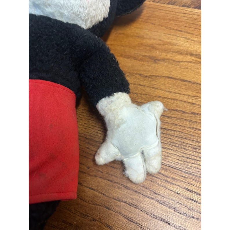 May include: A vintage Mickey Mouse soft toy, with a black body, red shorts, and white gloves. The toy has a classic design with signs of wear, indicating its age. The toy is placed on a wooden surface.