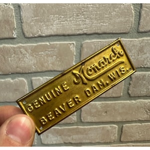 May include: A rectangular brass plate with the words "GENUINE Monarch BEAVER DAM, WIS." embossed on it. The plate has a vintage look with a slightly tarnished finish and small holes in each corner.