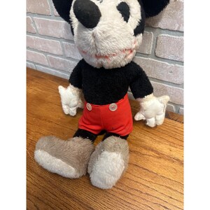 May include: A vintage Mickey Mouse soft toy. The toy has a black body, red shorts, white gloves, and large, fluffy, grey and white shoes. The face is white with black eyes and a black nose. The toy is on a wooden surface.