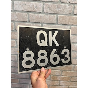 May include: A rectangular black metal license plate featuring the white alphanumeric characters QK 8863. The plate has a silver-toned border and two visible mounting holes positioned between the top and bottom rows of characters.