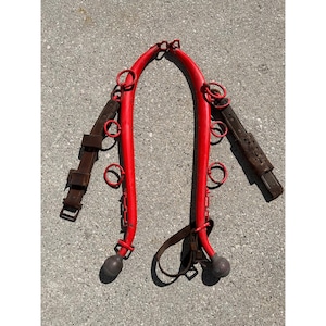 May include: A red horse collar with brown leather straps and metal rings. The collar has a curved shape with two rounded ends. The leather straps have buckles and are attached to the collar with metal rings.
