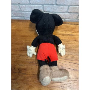 May include: A vintage Mickey Mouse soft toy, seen from the rear. It has a black head and body, red shorts, white gloves, and tan boots. The toy is placed on a wooden surface.
