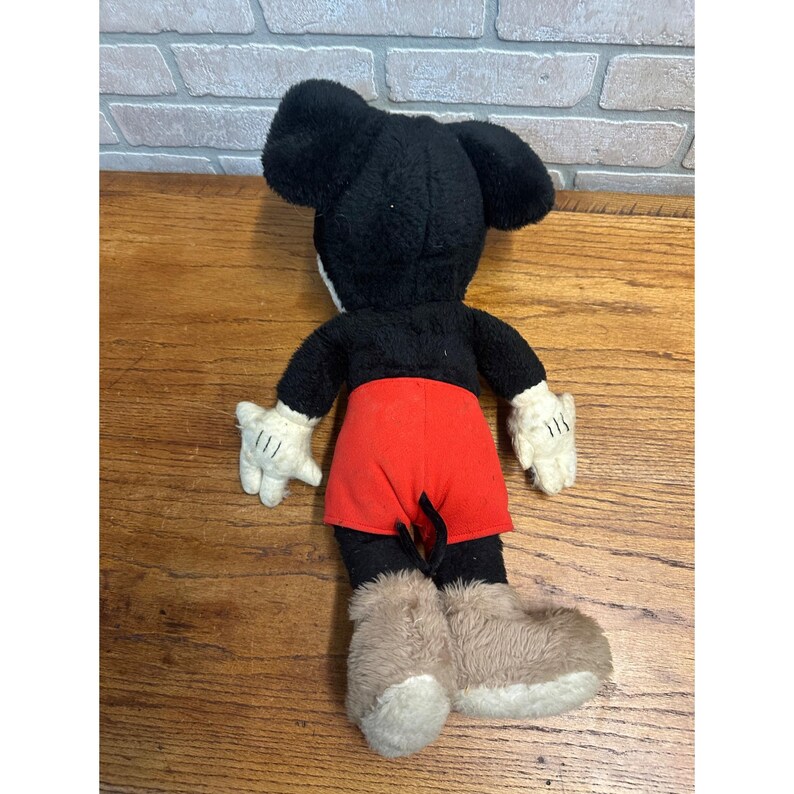 May include: A vintage Mickey Mouse soft toy, seen from the rear. It has a black head and body, red shorts, white gloves, and tan boots. The toy is placed on a wooden surface.