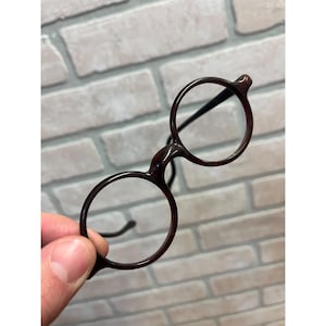 May include: A pair of round-framed glasses with a dark brown tortoiseshell pattern. The frames feature thin, circular rims and slender temples. The design is simple and functional, showcasing a classic circular shape with a smooth, polished finish.